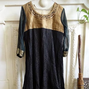 May include: A long, black dress with gold accents. The bodice is gold with intricate embroidery around the neckline. The sleeves are sheer, and the cuffs are trimmed with gold. The hemline also features a gold trim.
