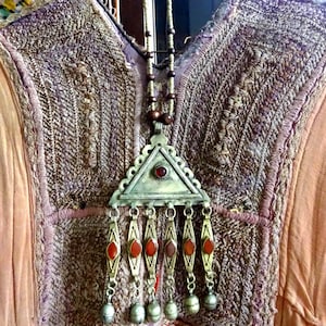 May include: A silver-toned pendant necklace with a triangular top and dangling teardrop-shaped charms, each with a red stone. The necklace hangs over a vintage, embroidered garment with floral fabric at the neckline. The garment is a mix of peach, purple, and brown.