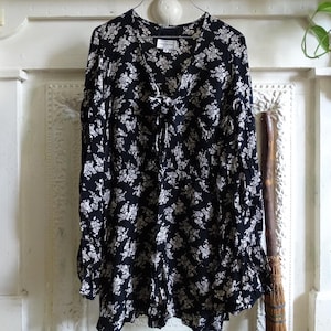 May include: Black floral print dress with a V-neckline and long sleeves. The dress features a tie detail at the neckline and a gathered waist. The fabric is patterned with small white flowers. The dress is hanging on a metal hanger.