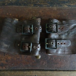 May include: Two brown leather wrist cuffs with three metal buckles each. The cuffs are worn and distressed, with visible stitching and metal hardware. The cuffs are likely vintage or antique.