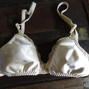 May include: A vintage, ivory-colored bra with a satin finish. The bra features delicate lace trim along the edges and adjustable straps. The cups are triangular in shape, and the overall design is classic and elegant.