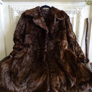 May include: A vintage brown fur coat with a classic design. The coat features a wide collar, button closures, and a knee-length hem. The fur appears to be in good condition, with a rich, dark brown color. The coat is hanging on a black hanger.