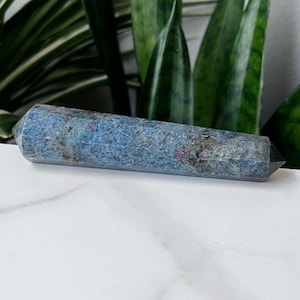 May include: A blue and gray crystal wand with a faceted, pointed end on both sides. The crystal has a speckled appearance with small red inclusions. The wand is displayed on a white surface with green foliage in the background.