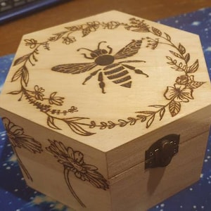 Può includere: Una scatola di legno esagonale con un coperchio incernierato e una chiusura in metallo. La scatola è decorata con un disegno di un'ape bruciata nel legno circondata da fiori e foglie.