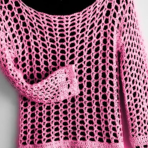 Könnte beinhalten: Ein leuchtend rosa Langarmpullover mit einem offenen Häkelmuster und einem weiten Rundhalsausschnitt. Das Kleidungsstück hat eine lockere, luftige Struktur mit einem festen, engmaschig gestrickten Saum am unteren Rand und an den Ärmelbündchen.