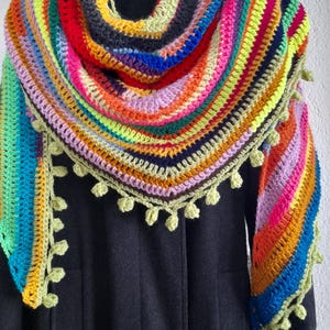 Peut inclure: Un châle crocheté à la main aux couleurs de l'arc-en-ciel, avec des nuances de rouge, orange, jaune, vert, bleu et violet. Le châle triangulaire présente un bord festonné et de petites décorations vertes en forme de feuilles.
