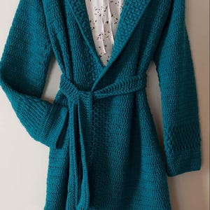 Könnte beinhalten: Ein tealblauer Häkelcardigan mit Kapuze und Bindegürtel. Der Cardigan hat ein strukturiertes Stichmuster und eine lange, fließende Silhouette.