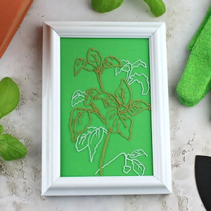May include: A white frame with a green background and a gold and white paper cutout of a basil plant.
