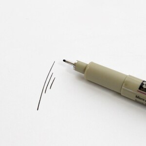 Op de afbeelding: Een zwarte fijnlijnstift met een beige cilinder en de tekst "Sakura Micro Pen" gedrukt op de zijkant.