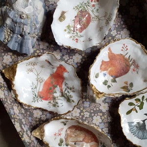 Könnte beinhalten: Sechs dekorative Austernschalen mit goldener Verzierung, jede mit einem anderen Waldtier und einem floralen Design bemalt. Die Tiere umfassen einen Fuchs, ein Eichhörnchen, ein Reh und einen Vogel.