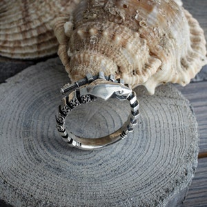Op de afbeelding: Een zilveren ring met een hartvormig ontwerp en ingewikkelde details. De ring wordt gepresenteerd op een houten schijf met een zeeschelp op de achtergrond. De band van de ring heeft een herhalend patroon.
