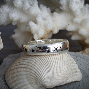 May include: A silver ring with star and moon cutouts. The ring is displayed on a seashell, with a white coral and other seashells in the background. The ring has a polished finish and a band width of approximately 0.25 inches.
