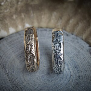 May include: Two textured metal rings, one gold-toned and one silver-toned, rest on a gray wooden surface. The rings feature an organic, patterned design. The background includes a blurred, neutral-toned element.