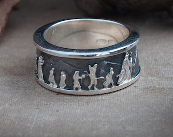 May include: A silver ring with a detailed relief design. The ring features a mountain range and figures in silhouette. The ring's band is wide, and the design wraps around the entire circumference. The ring is set against a neutral background.