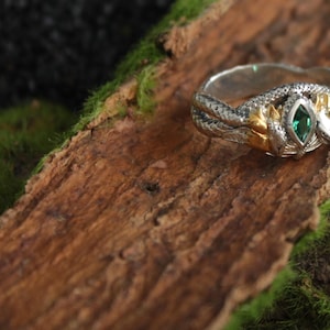 K&ouml;nnte beinhalten: Ein silberner und goldener Ring mit Schlangenmuster. Der Ring hat einen gr&uuml;nen Edelstein in der Mitte und liegt auf einem Holzst&uuml;ck mit Moos. Der Ring hat aufwendige Details und einen einzigartigen Stil.