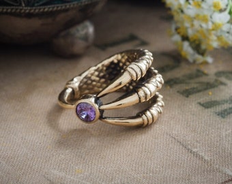 Op de afbeelding: Een goudkleurige ring met een klauwontwerp, met drie gebogen tanden en een centrale paarse edelsteen. De ring is geplaatst tegen een neutrale achtergrond met witte bloemen en een decoratieve kom.