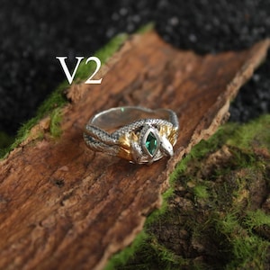 K&ouml;nnte beinhalten: Ein silberner und goldener Ring mit Schlangenmuster und einem gr&uuml;nen Edelstein. Der Ring liegt auf einem Holzst&uuml;ck mit Moos, mit dem Text "V2" sichtbar.