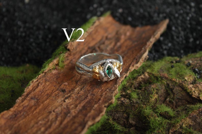K&ouml;nnte beinhalten: Ein silberner und goldener Ring mit Schlangenmuster und einem gr&uuml;nen Edelstein. Der Ring liegt auf einem Holzst&uuml;ck mit Moos, mit dem Text "V2" sichtbar.