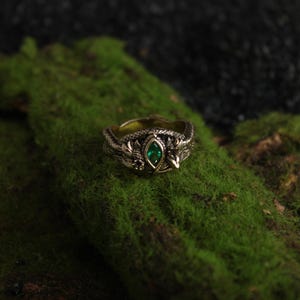 K&ouml;nnte beinhalten: Ein detaillierter goldfarbener Ring mit einem ovalen, smaragdgr&uuml;nen Edelstein in der Mitte. Der Ring hat ein verziertes Design mit Blatt- und drachenartigen Details. Der Ring ist vor einem Hintergrund aus gr&uuml;nem Moos platziert.