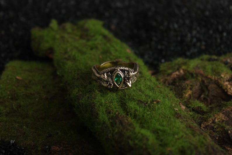 K&ouml;nnte beinhalten: Ein detaillierter goldfarbener Ring mit einem ovalen, smaragdgr&uuml;nen Edelstein in der Mitte. Der Ring hat ein verziertes Design mit Blatt- und drachenartigen Details. Der Ring ist vor einem Hintergrund aus gr&uuml;nem Moos platziert.