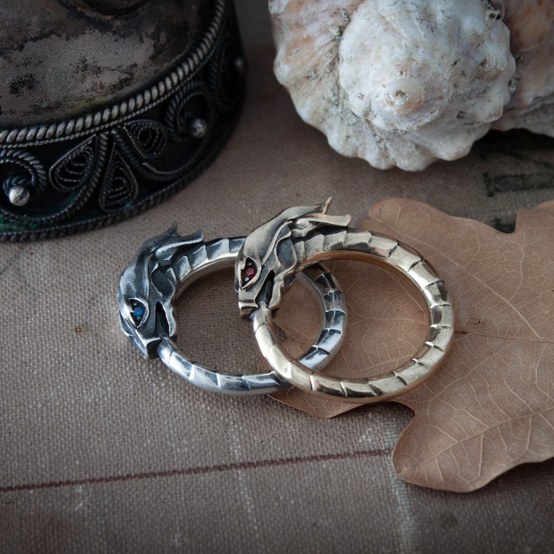 Puede incluir: Dos anillos con cabeza de drag&oacute;n, uno plateado con un ojo de gema azul, el otro dorado con un ojo de gema roja. Los anillos son circulares y detallados, con las cabezas de drag&oacute;n enfrentadas. Los anillos est&aacute;n sobre una superficie texturizada.