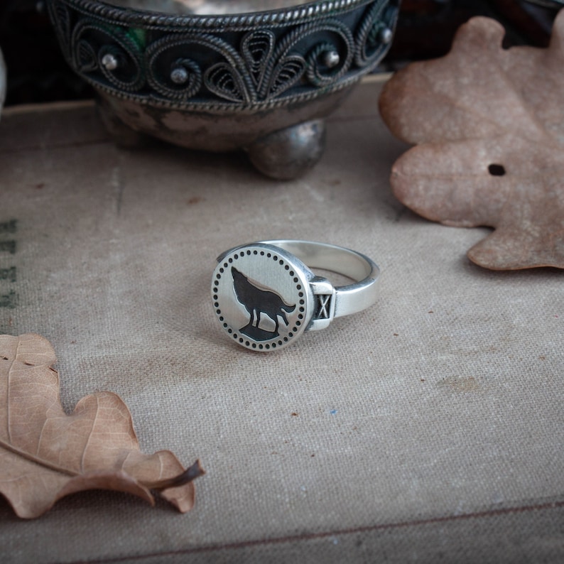 May include: A silver ring with a circular face featuring a black wolf howling at the moon. The ring has a polished finish and a small rectangular detail with an "X" on the side. The ring is set against a neutral background with a leaf.
