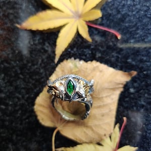 K&ouml;nnte beinhalten: Ein silberner Ring mit einem gr&uuml;nen Edelstein in der Mitte, auf einem braunen Blatt. Der Ring hat aufwendige Details mit goldenen Akzenten. Der Hintergrund ist eine dunkle, strukturierte Oberfl&auml;che mit gelben Bl&auml;ttern.