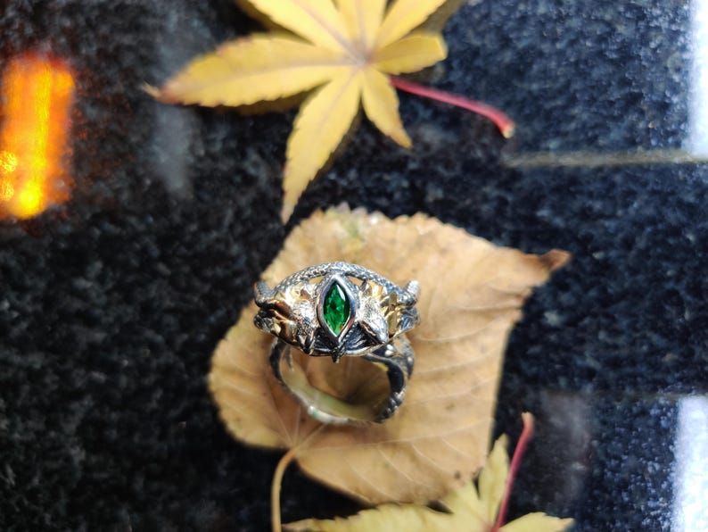 K&ouml;nnte beinhalten: Ein silberner Ring mit einem gr&uuml;nen Edelstein in der Mitte, auf einem braunen Blatt. Der Ring hat aufwendige Details mit goldenen Akzenten. Der Hintergrund ist eine dunkle, strukturierte Oberfl&auml;che mit gelben Bl&auml;ttern.