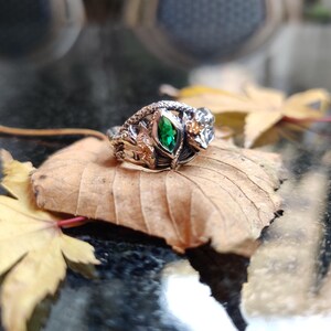 K&ouml;nnte beinhalten: Ein detaillierter Silberring mit einem zentralen, smaragdgeschliffenen gr&uuml;nen Edelstein. Der Ring weist komplizierte Blatt- und Rankenmuster mit zus&auml;tzlichen kleinen Edelsteinen auf. Er liegt auf einem getrockneten Blatt, mit weiteren Bl&auml;ttern im Hintergrund.
