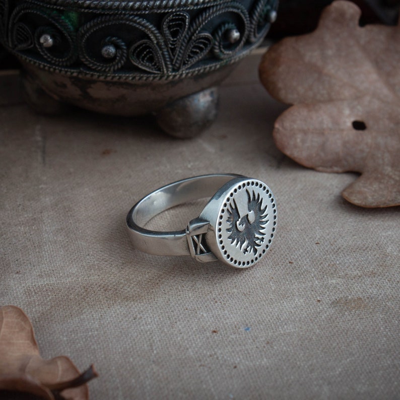 May include: A silver ring with a circular face featuring a detailed black bird design. The ring has a band with an 'X' marking on the side. The ring is displayed on a neutral-toned surface.