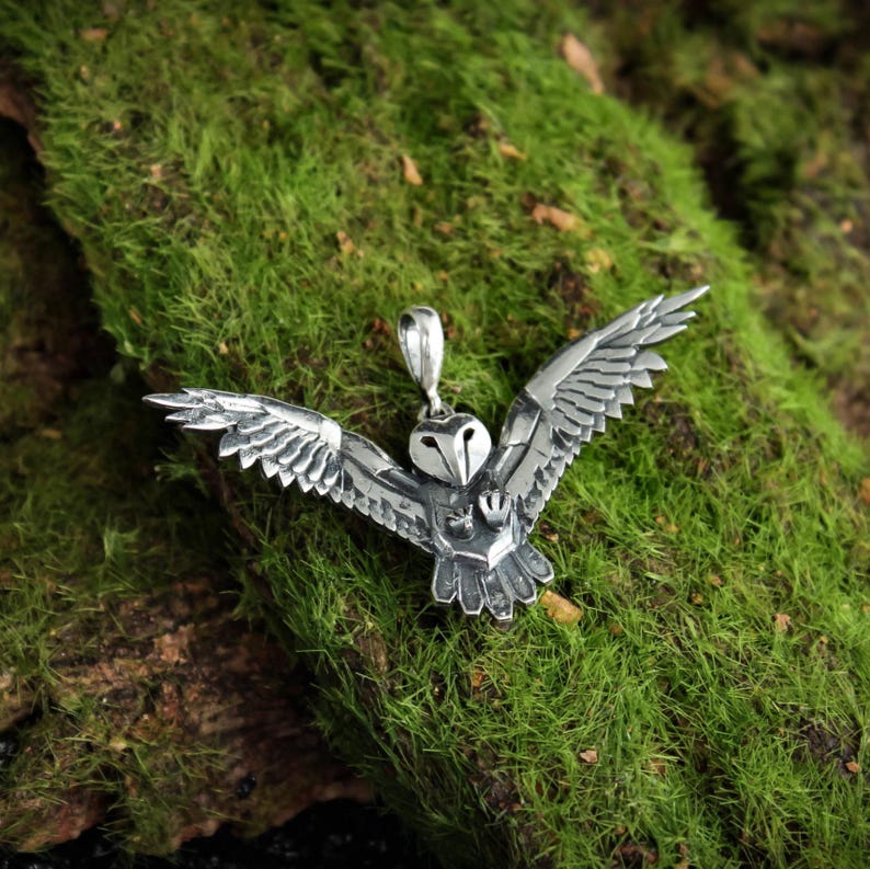 May include: A detailed silver owl pendant with outstretched wings. The pendant is intricately designed with textured feathers and a small loop for a chain. The owl is centered against a backdrop of green moss.
