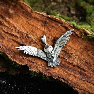 May include: A silver-colored owl pendant with outstretched wings. The pendant is detailed with feathers and a small loop for a chain. The owl is centered on a piece of brown wood with green moss.