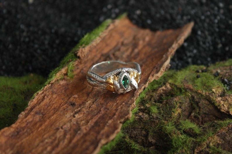 K&ouml;nnte beinhalten: Ein silberner und goldener Ring mit Schlangenmuster. Der Ring hat einen gr&uuml;nen Edelstein in der Mitte und liegt auf einem Holzst&uuml;ck mit Moos. Der Ring hat aufwendige Details und einen einzigartigen Stil.