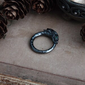 Puede incluir: Un anillo gris oscuro con un dise&ntilde;o de cabeza de drag&oacute;n. La cabeza del drag&oacute;n presenta ojos de piedra preciosa verde. El anillo est&aacute; sobre un fondo neutro con pi&ntilde;as.