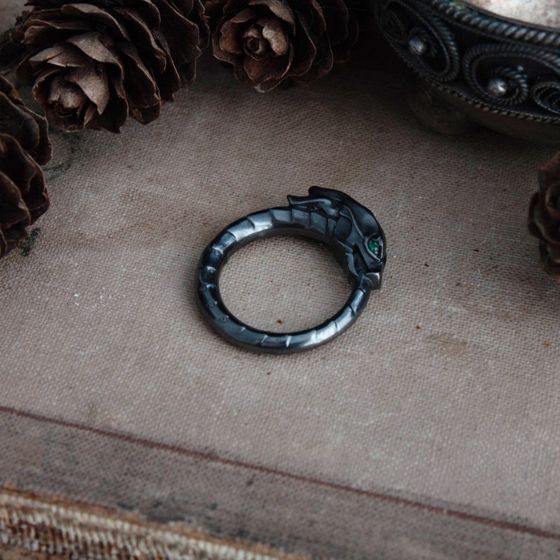 Puede incluir: Un anillo gris oscuro con un dise&ntilde;o de cabeza de drag&oacute;n. La cabeza del drag&oacute;n presenta ojos de piedra preciosa verde. El anillo est&aacute; sobre un fondo neutro con pi&ntilde;as.