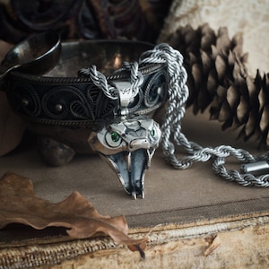 May include: A silver cobra pendant necklace with green gemstone eyes. The snake's mouth is open, revealing its fangs. The necklace has a thick, braided chain. The pendant is detailed and sits on a vintage book.