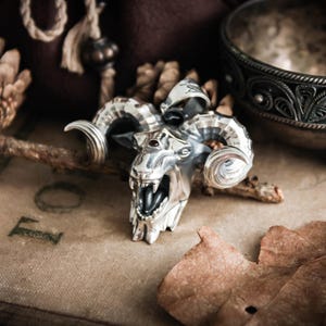 May include: A detailed silver pendant in the shape of a ram's head, featuring curved horns, an open mouth with visible teeth, and a small red gem. The pendant is set against a backdrop of dried leaves and a vintage-style surface.