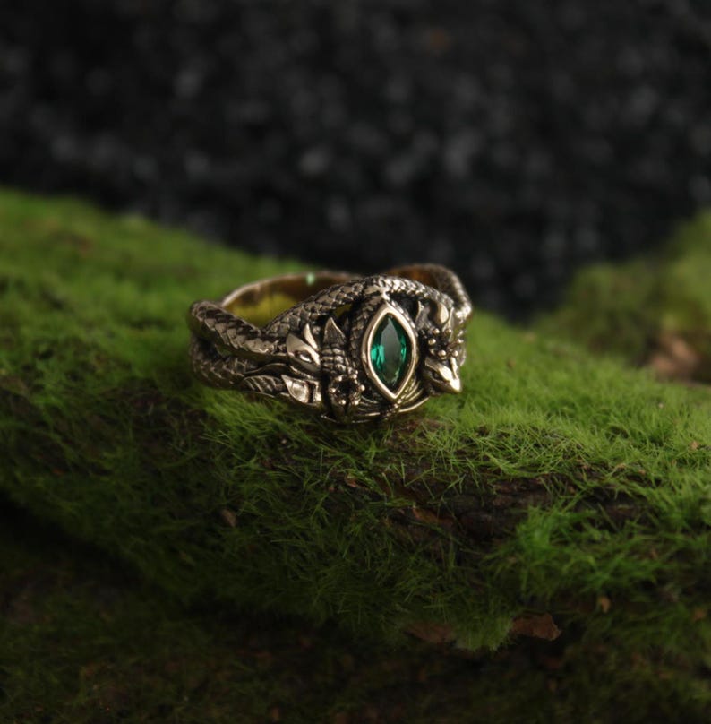 K&ouml;nnte beinhalten: Ein detaillierter Ring im antiken Stil mit Schlangenmotiv. Der Ring ist bronzefarben mit aufwendigen Schnitzereien und einem zentralen, smaragdgr&uuml;nen Edelstein. Pr&auml;sentiert auf gr&uuml;nem Moos.