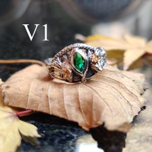 K&ouml;nnte beinhalten: Ein detaillierter Silberring mit einem zentralen smaragdgr&uuml;nen Edelstein. Der Ring weist aufwendige Designs auf, darunter eine Schlange und florale Elemente. Der Text "V1" ist sichtbar. Der Ring liegt auf einem braunen Blatt.