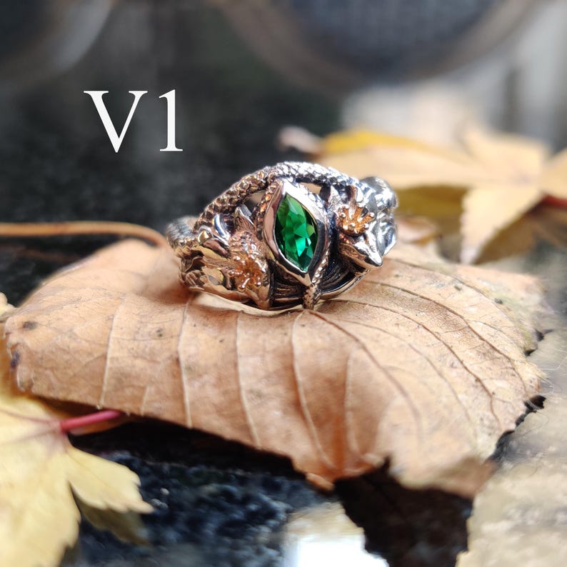 K&ouml;nnte beinhalten: Ein detaillierter Silberring mit einem zentralen smaragdgr&uuml;nen Edelstein. Der Ring weist aufwendige Designs auf, darunter eine Schlange und florale Elemente. Der Text "V1" ist sichtbar. Der Ring liegt auf einem braunen Blatt.