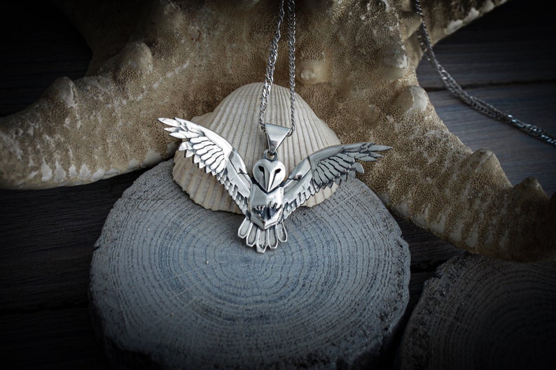 May include: A silver-colored owl pendant with outstretched wings, suspended from a delicate chain. The owl's detailed feathers and facial features are visible. The pendant rests on a seashell and a wooden surface.