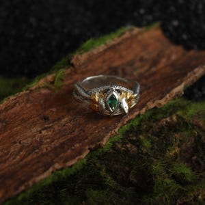 K&ouml;nnte beinhalten: Ein silberner und goldener Ring mit Schlangenmuster, mit einem zentralen smaragdgr&uuml;nen Edelstein. Der Ring liegt auf einem Holzst&uuml;ck mit Moos, was eine nat&uuml;rliche und k&uuml;nstlerische Pr&auml;sentation ergibt. Der Schlangenkopf ist sichtbar.