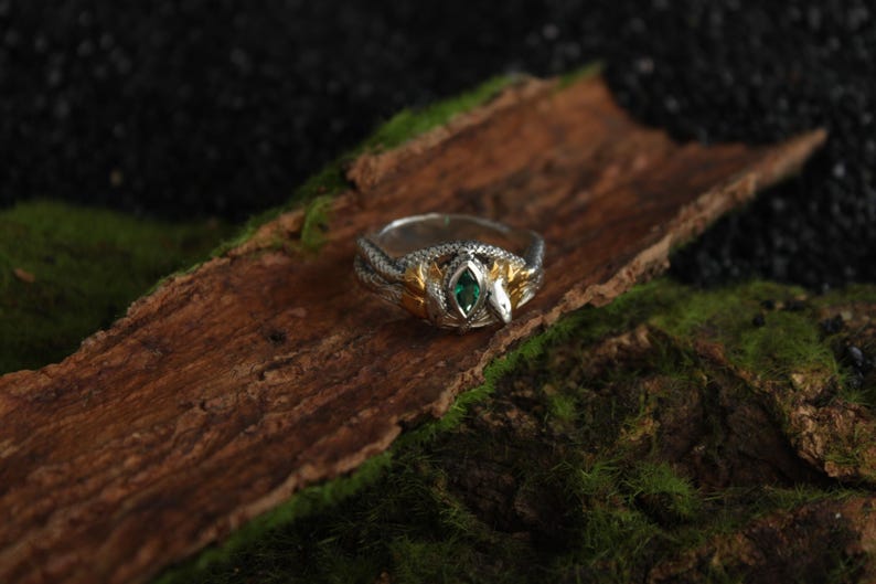 K&ouml;nnte beinhalten: Ein silberner und goldener Ring mit Schlangenmuster, mit einem zentralen smaragdgr&uuml;nen Edelstein. Der Ring liegt auf einem Holzst&uuml;ck mit Moos, was eine nat&uuml;rliche und k&uuml;nstlerische Pr&auml;sentation ergibt. Der Schlangenkopf ist sichtbar.