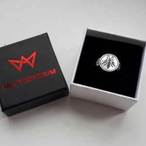 May include: A silver ring with a bee design inside a circle, presented in a white box with a black interior. The ring is next to a black box with the word "MASTENARIUM" in red.