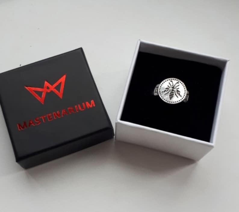 May include: A silver ring with a bee design inside a circle, presented in a white box with a black interior. The ring is next to a black box with the word "MASTENARIUM" in red.