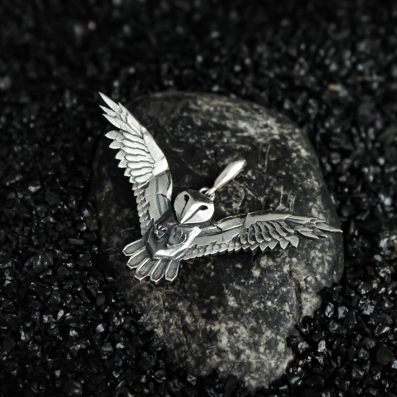 May include: A silver-colored owl pendant with outstretched wings, resting on a dark gray stone. The owl's wings and body are intricately detailed, showcasing a textured surface. The pendant is designed to appear as if the owl is in flight.