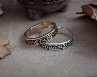 May include: Two rings with an ornate, swirling pattern. One ring is gold-toned, the other is silver-toned. The rings are set on a neutral-colored surface, with a decorative metal container in the background.