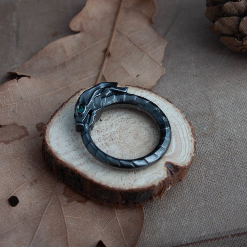 Puede incluir: Anillo de drag&oacute;n gris oscuro con un ojo de gema verde. El anillo de metal est&aacute; colocado sobre una peque&ntilde;a rodaja de madera. La cabeza del drag&oacute;n est&aacute; detallada con escamas y una superficie texturizada.