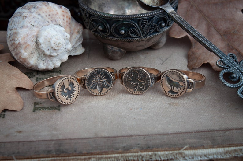 May include: Four antique-style rings with bronze bands and circular faces. Each ring features a different embossed design: a bird, a bee, a lion, and a wolf. The rings are displayed on a vintage book.