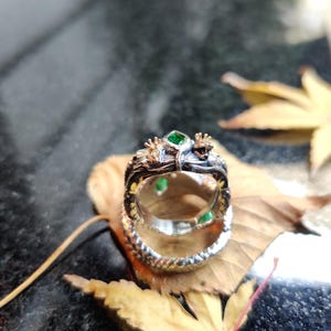 K&ouml;nnte beinhalten: Ein detaillierter Silberring mit einem Drachendesign, der gr&uuml;ne Edelsteine ​​aufweist. Der Kopf und der K&ouml;rper des Drachen umschlie&szlig;en den Ring, wobei die Edelsteine ​​in das Design eingesetzt sind. Der Ring wird auf einem Blatt pr&auml;sentiert.